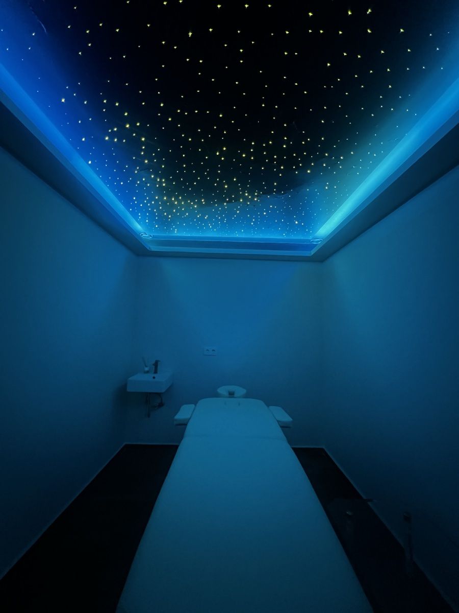Sala de masajes con techo estrellado e iluminación azul. Una camilla vacía en el centro.