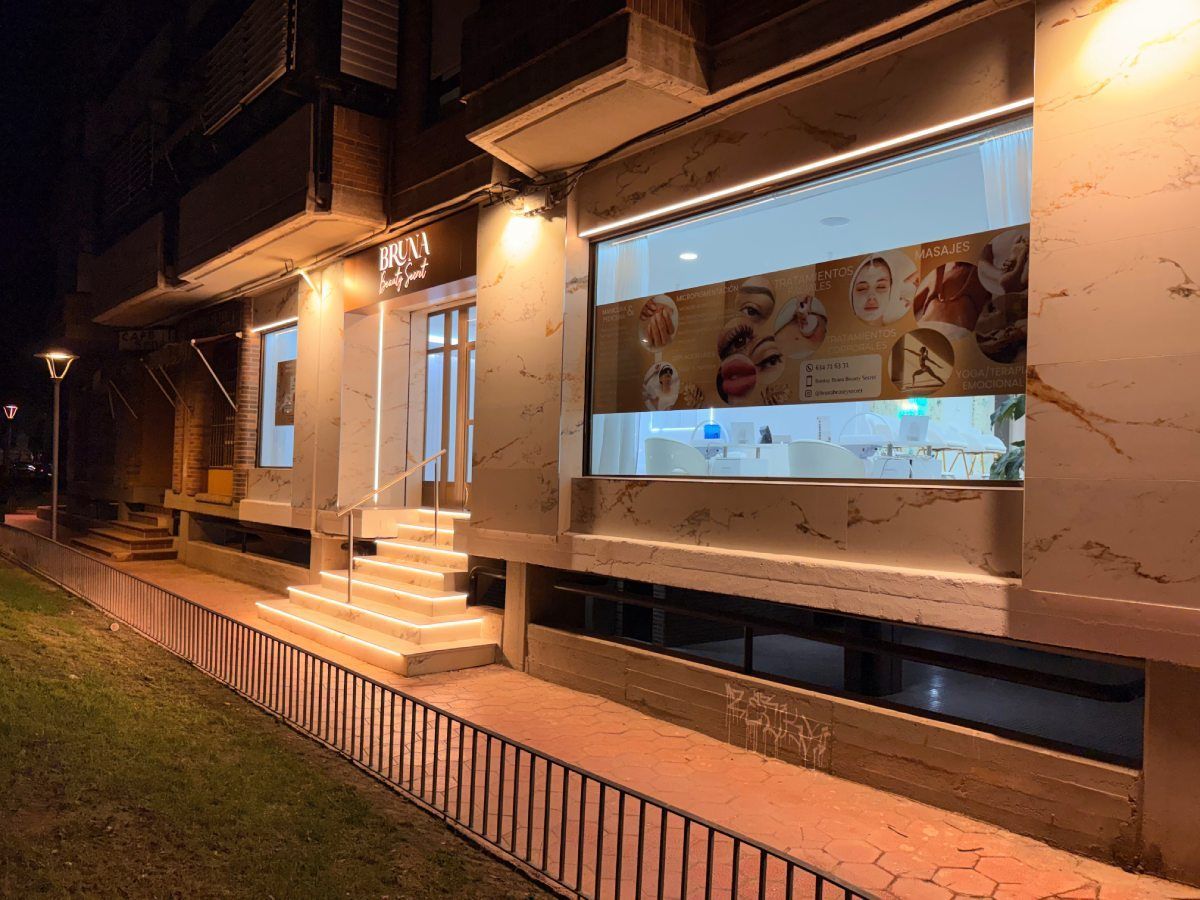 Fachada del edificio con entrada iluminada, escaleras y un gran ventanal con imágenes promocionales. De noche.
