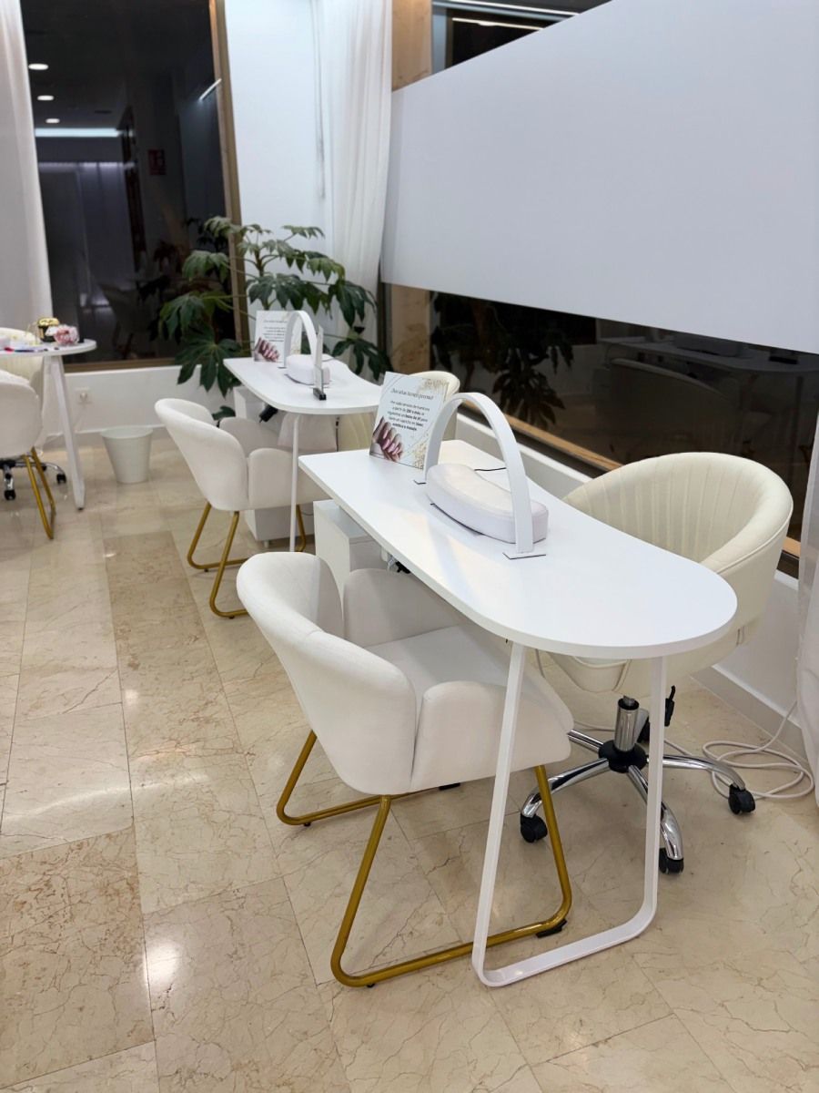 Interior de salón de uñas con mesas y sillas blancas, detalles dorados y una ventana de vidrio esmerilado.