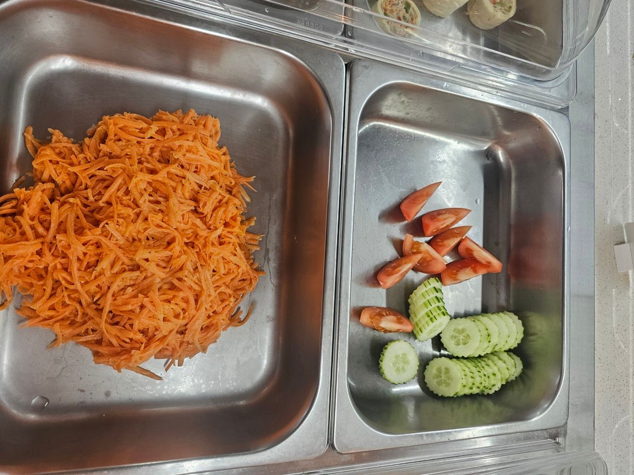 Bacs de service en métal contenant des carottes râpées, des tomates tranchées et des rondelles de concombre.