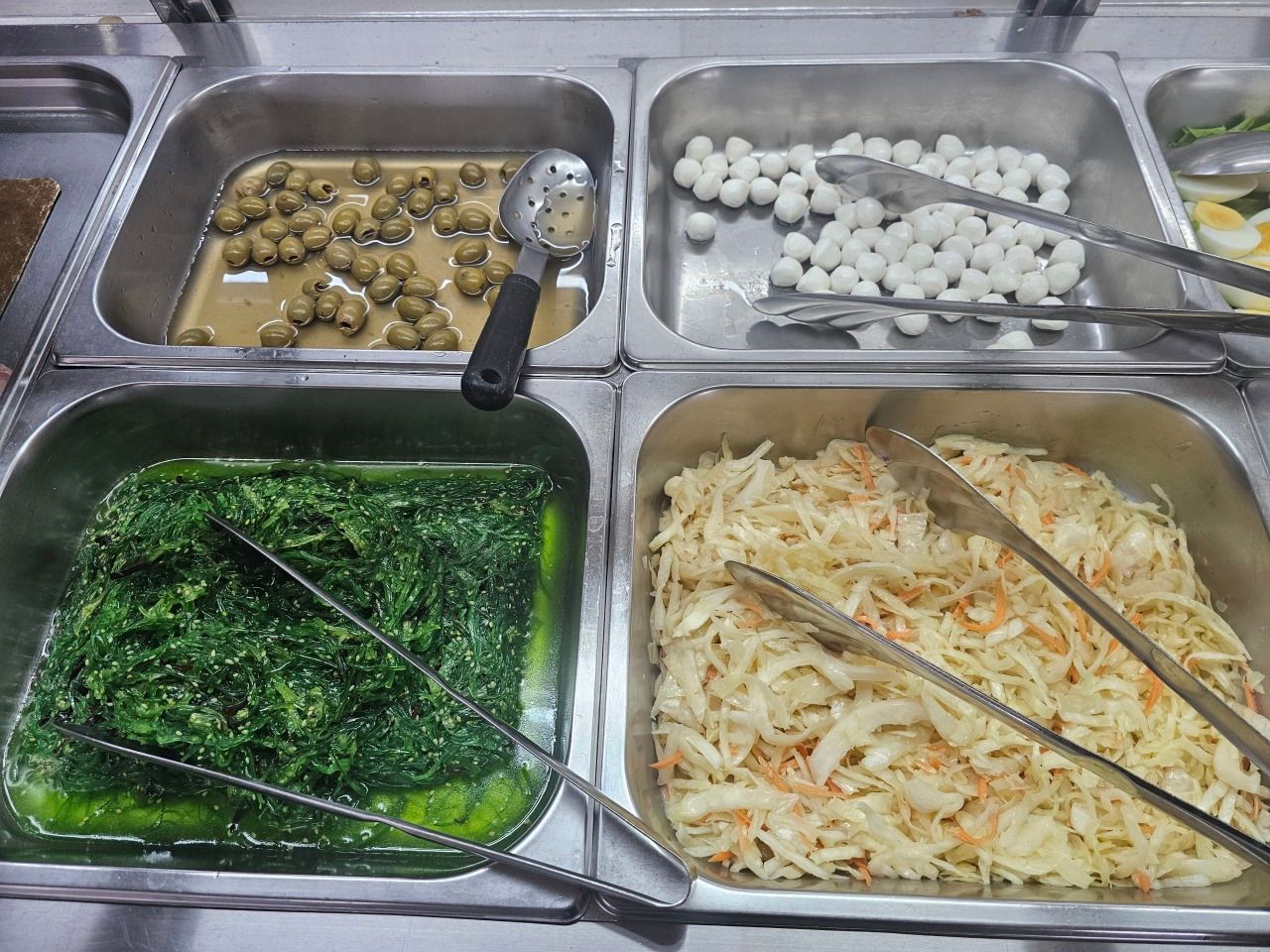 Quatre récipients contenant des olives, des billes de mozzarella, de la salade d'algues et de la salade de carottes râpées.