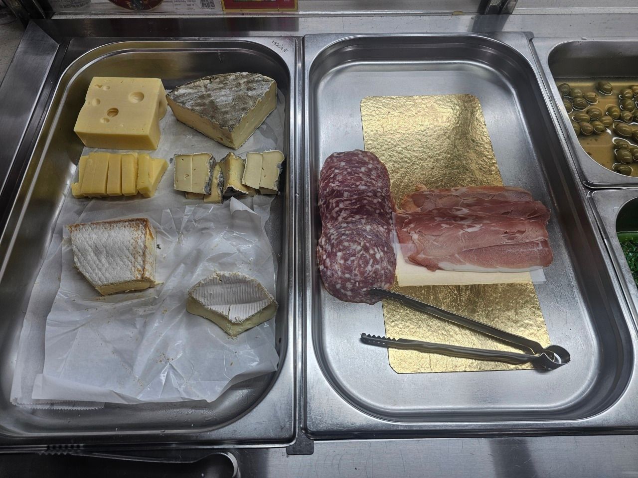 Deux plateaux avec divers fromages tranchés, du salami et de la charcuterie, accompagnés d'une paire de pinces métalliques.