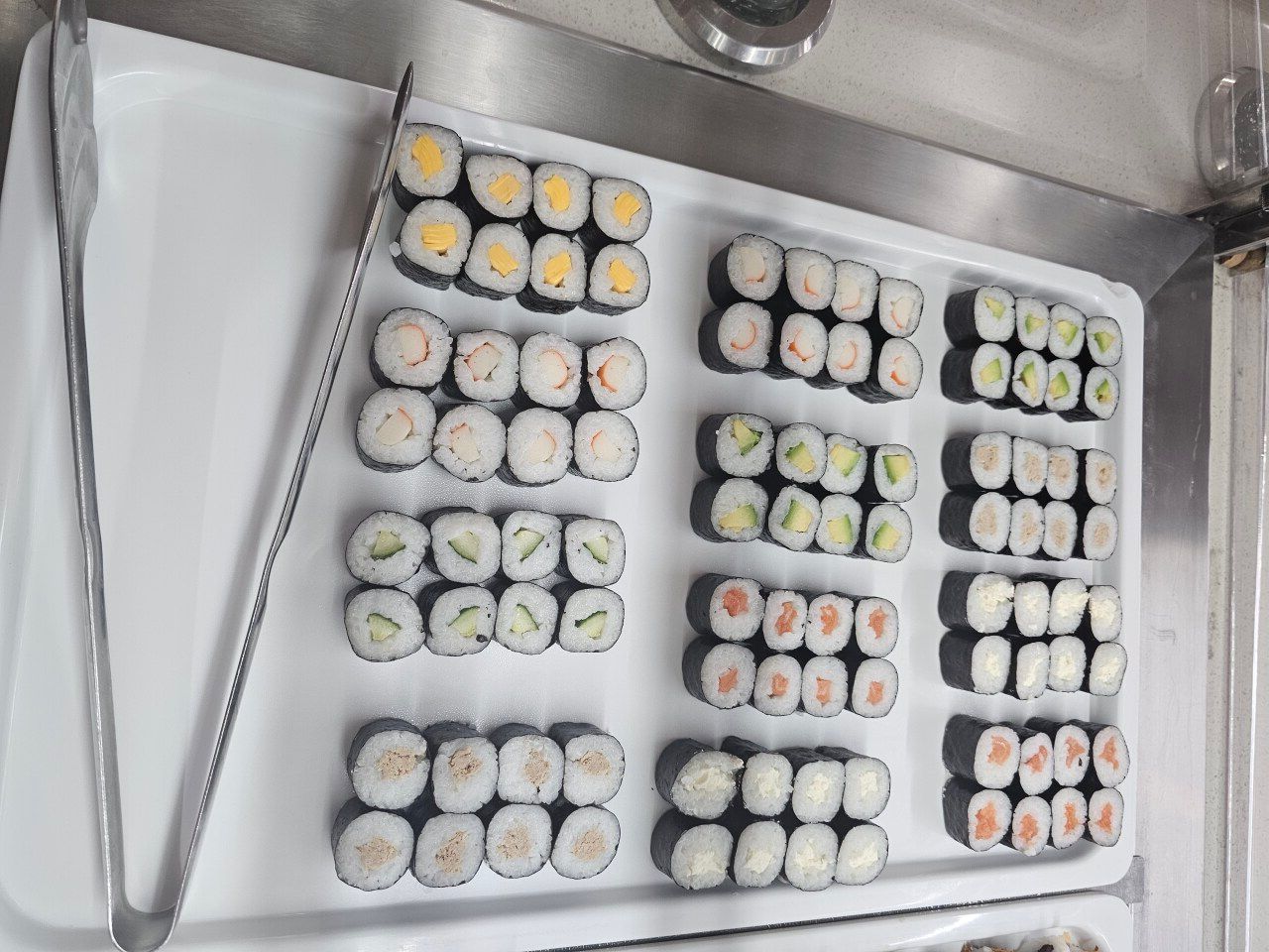 Un plateau de service présentant plusieurs rangées de sushis, accompagné de pinces métalliques sur le côté gauche.