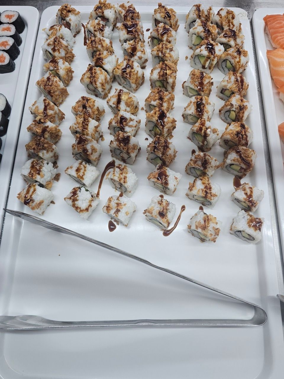 Un plat présentant des rangées de rouleaux de sushis nappés de sauce foncée, placé à côté d'une pince de service en métal.
