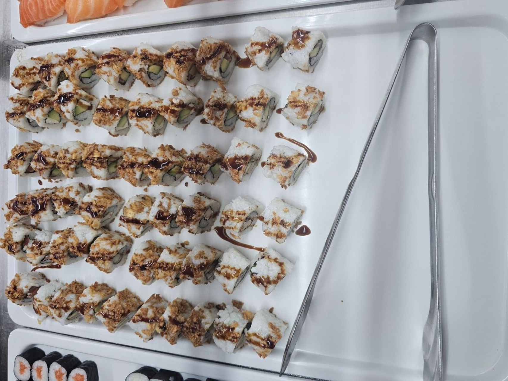 Vue de dessus d'un plateau blanc rempli de rouleaux de sushi nappés de sauce foncée et d'une paire de pinces en métal.