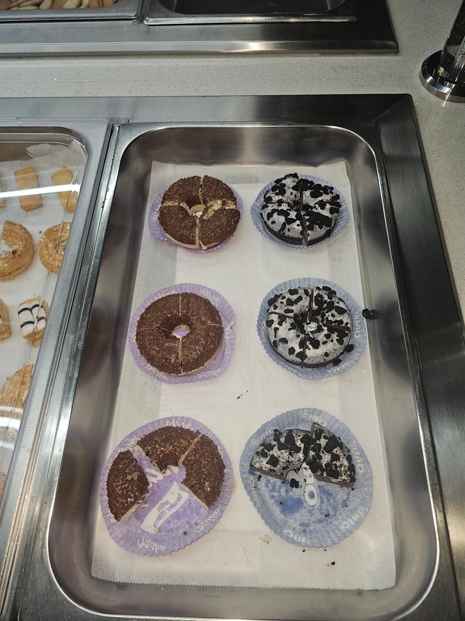 Six beignets, garnis de chocolat et de biscuits, disposés dans un plateau métallique de cafétéria.