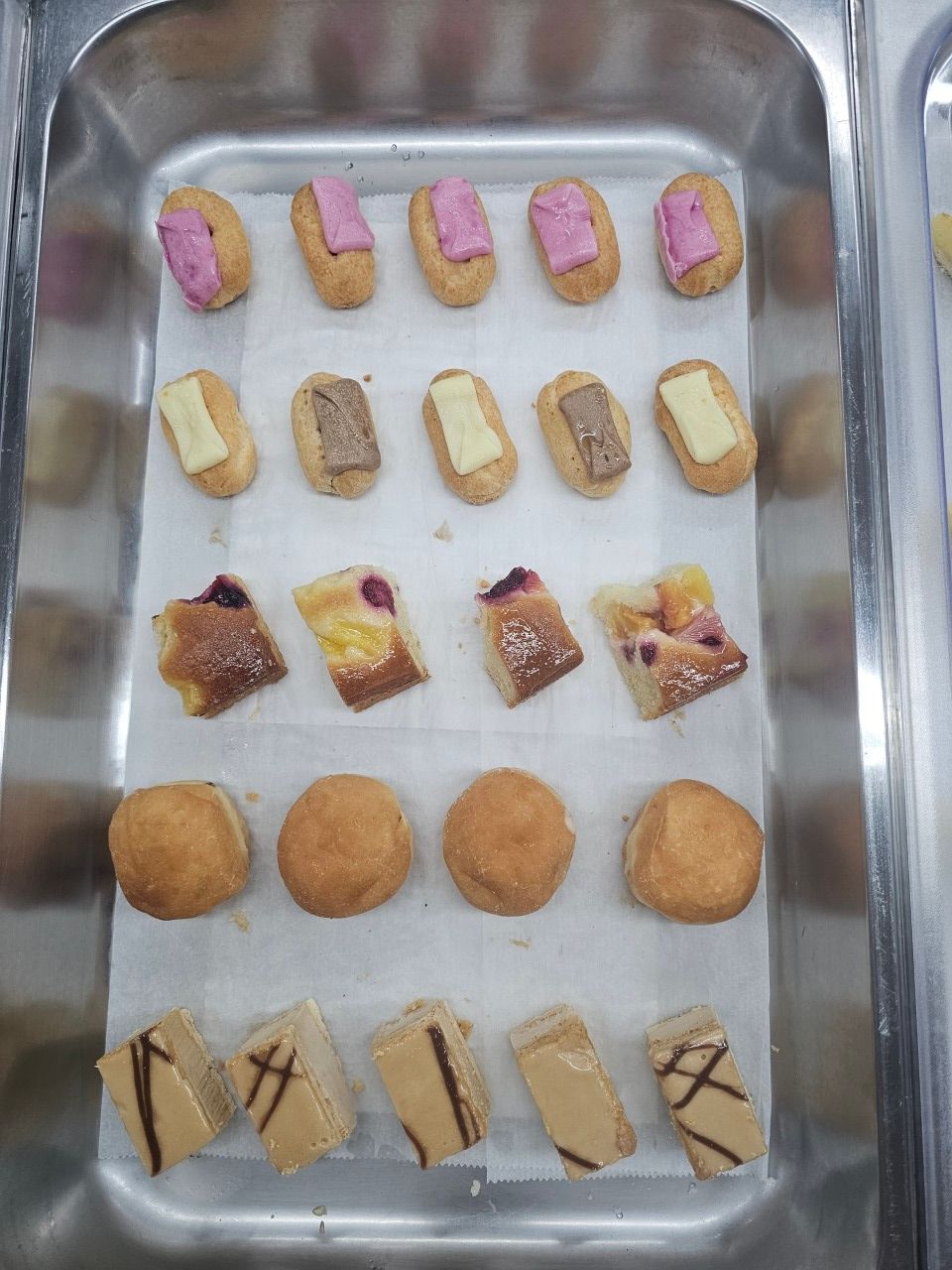 Un plateau contenant cinq rangées de pâtisseries variées, dont des éclairs glacés, des gâteaux et des petits pains ronds.