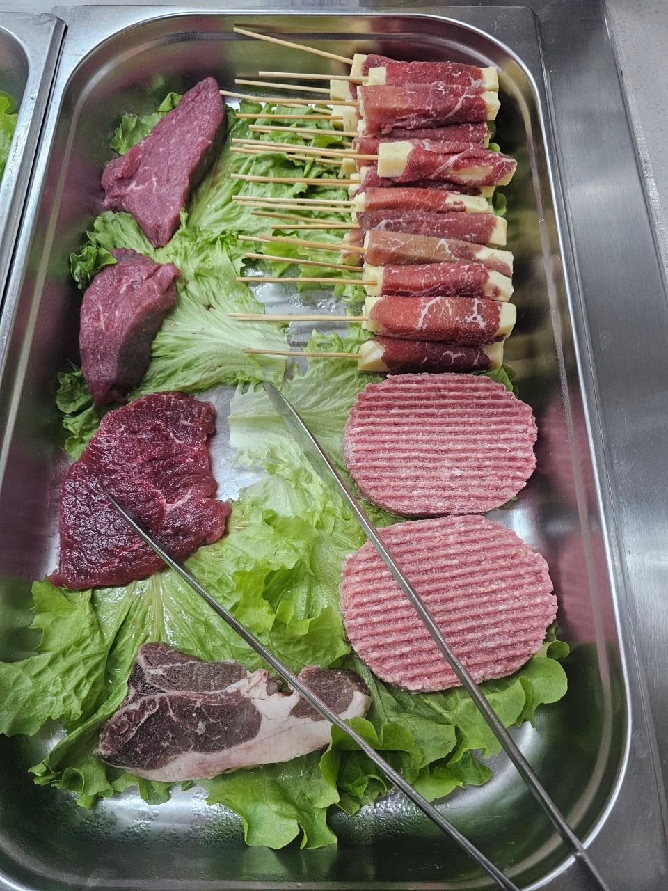 Un plateau en acier inoxydable contient des steaks crus, deux steaks hachés et des brochettes de viande et de fromage.