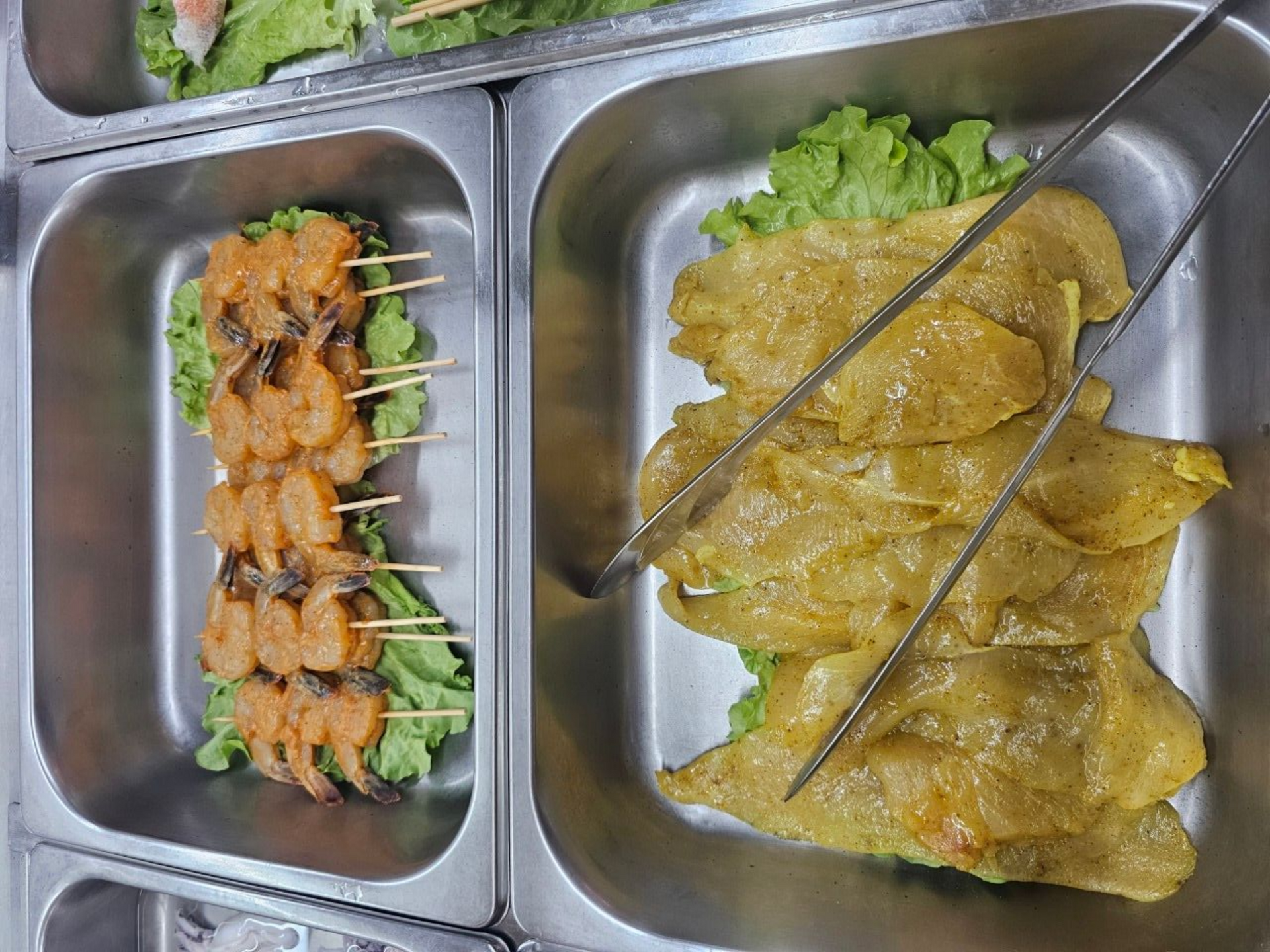 Plateaux en acier contenant des brochettes de crevettes et des filets de poulet jaune marinés, avec pinces de service.