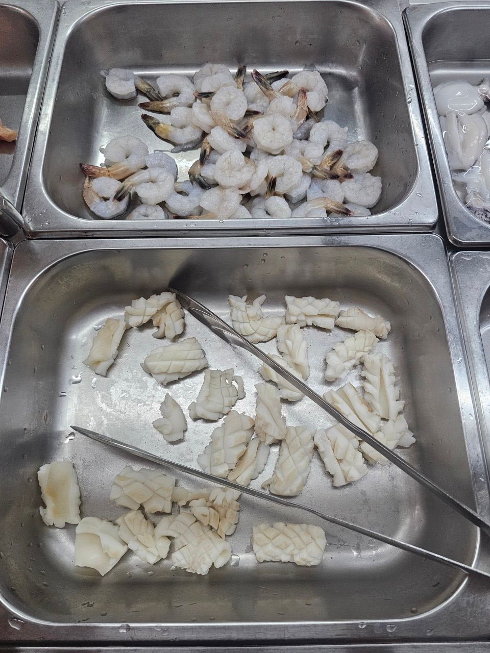 Deux barquettes dans une vitrine réfrigérée : l'une contient des crevettes crues, l'autre des morceaux de calamars incisés.