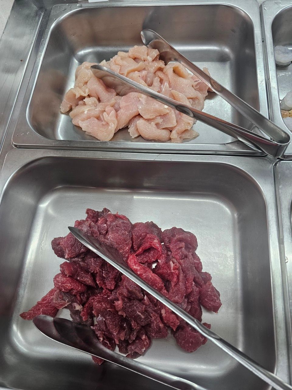 Deux plats métalliques disposés sur un buffet, l'un contenant des lanières de poulet cru et l'autre des morceaux de bœuf cru.