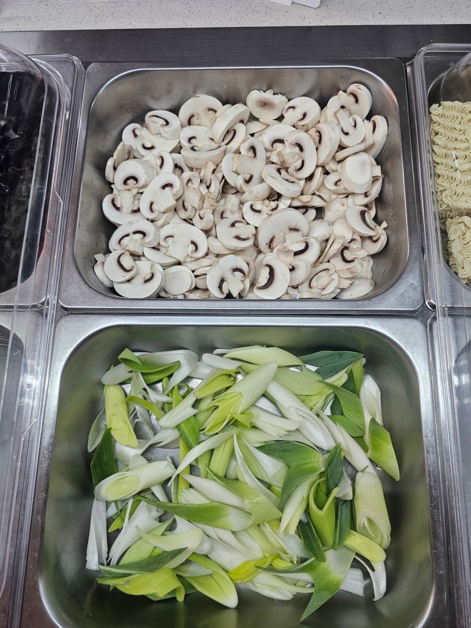 Deux récipients métalliques dans un buffet, l'un rempli de champignons blancs émincés et l'autre de poireaux verts hachés.