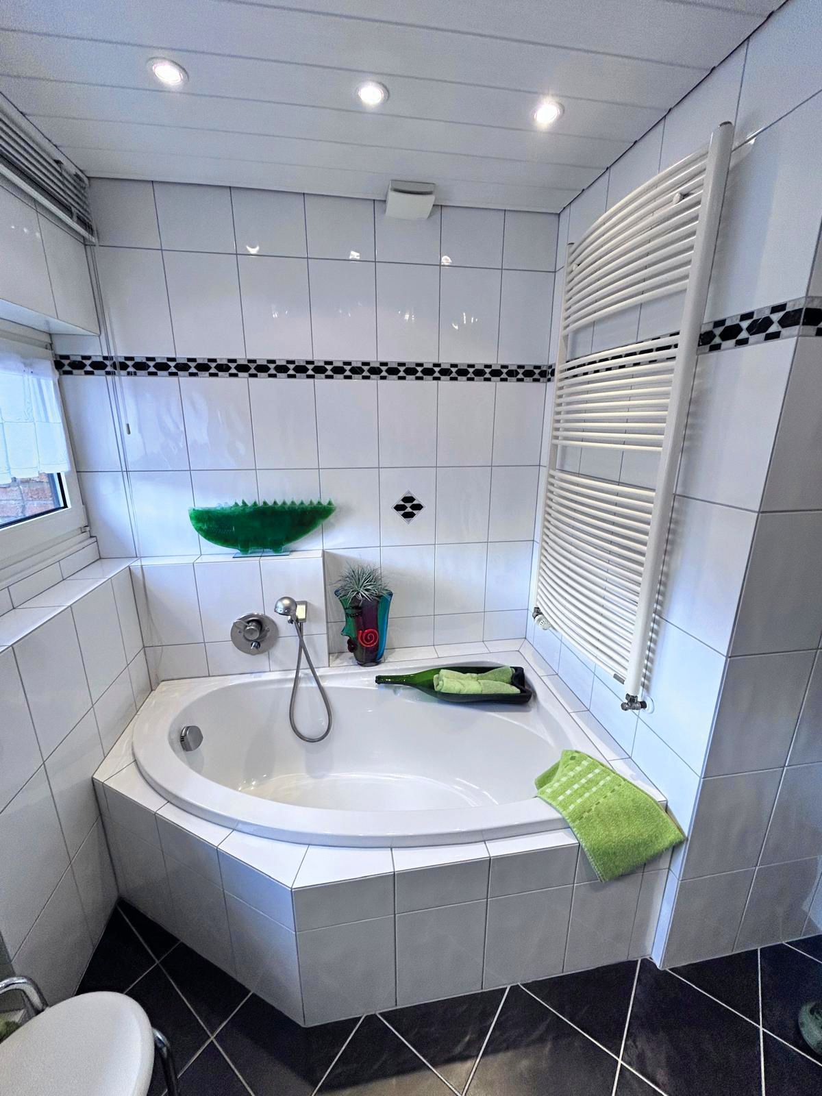 Helles, weißes Badezimmer mit Eckbadewanne, Handbrause, grünen Akzenten und schwarzem Fliesenboden