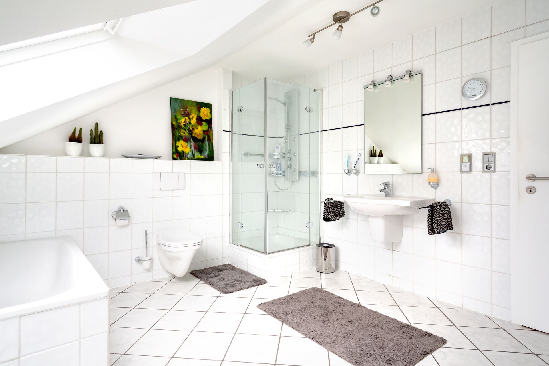 Helles, weißes Badezimmer mit Badewanne, gefliesten Wänden, Dusche, Waschbecken, Spiegel und grauen Badematten.