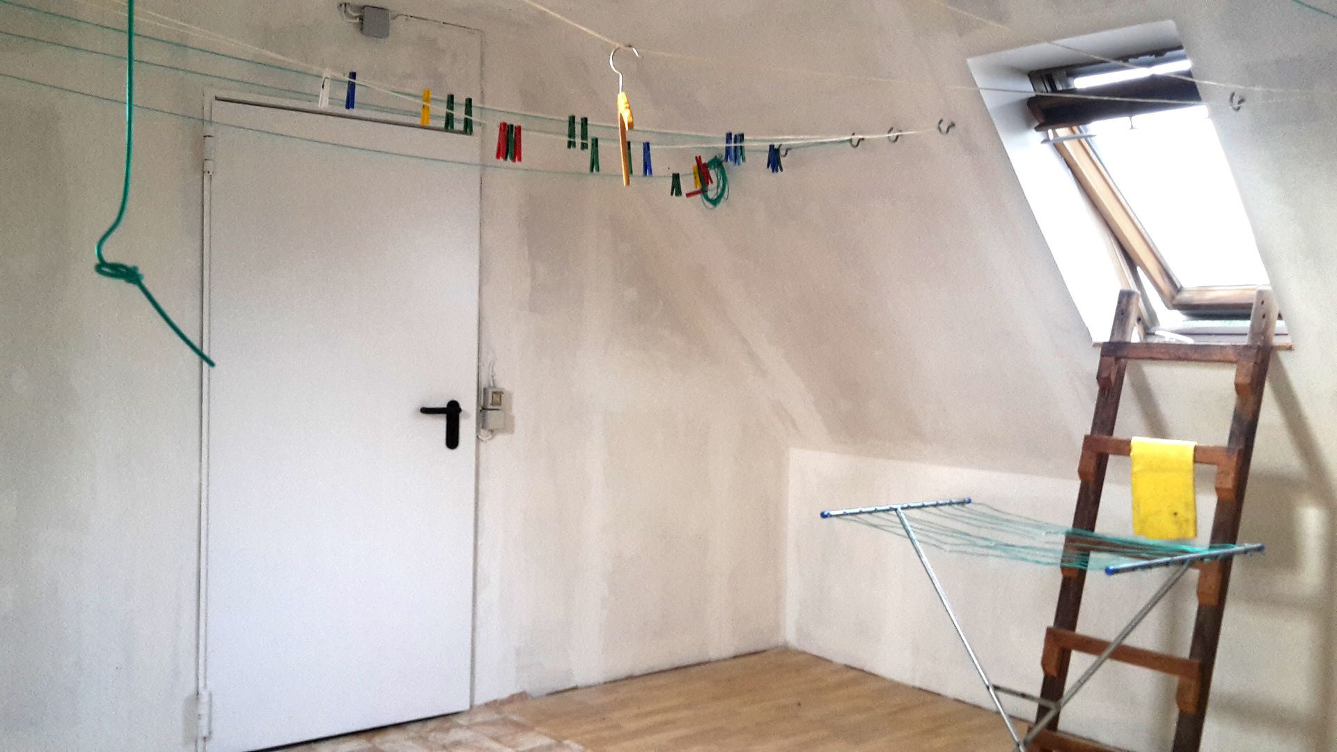 Kleiner Dachbodenraum mit weißer Tür, hängenden Wäscheklammern und einem Wäscheständer neben einem Dachfenster.