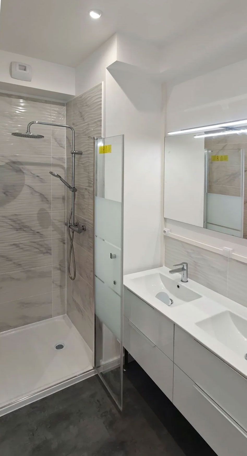 Salle de bains moderne avec douche et meuble-lavabo double vasque. Tons blancs et gris, design épuré.