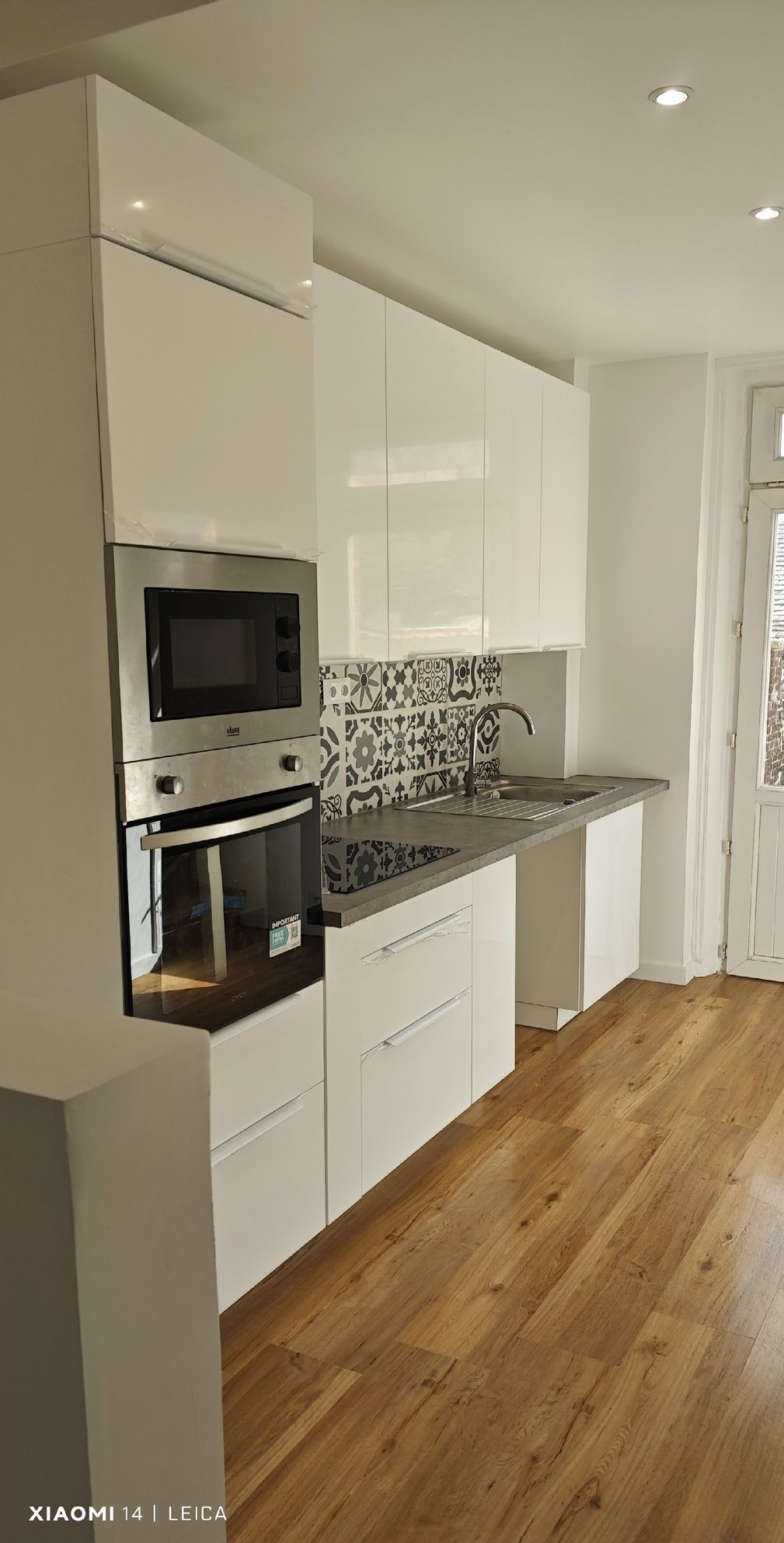 Cuisine blanche avec four encastré, micro-ondes et placards. Évier en inox et crédence noire et blanche. Sol en bois.