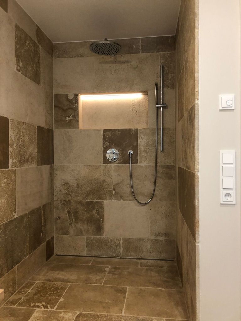 Dusche mit Steinfliesen in Hellbraun- und Brauntönen, Einbauregal mit warmer Beleuchtung und silbernen Armaturen.