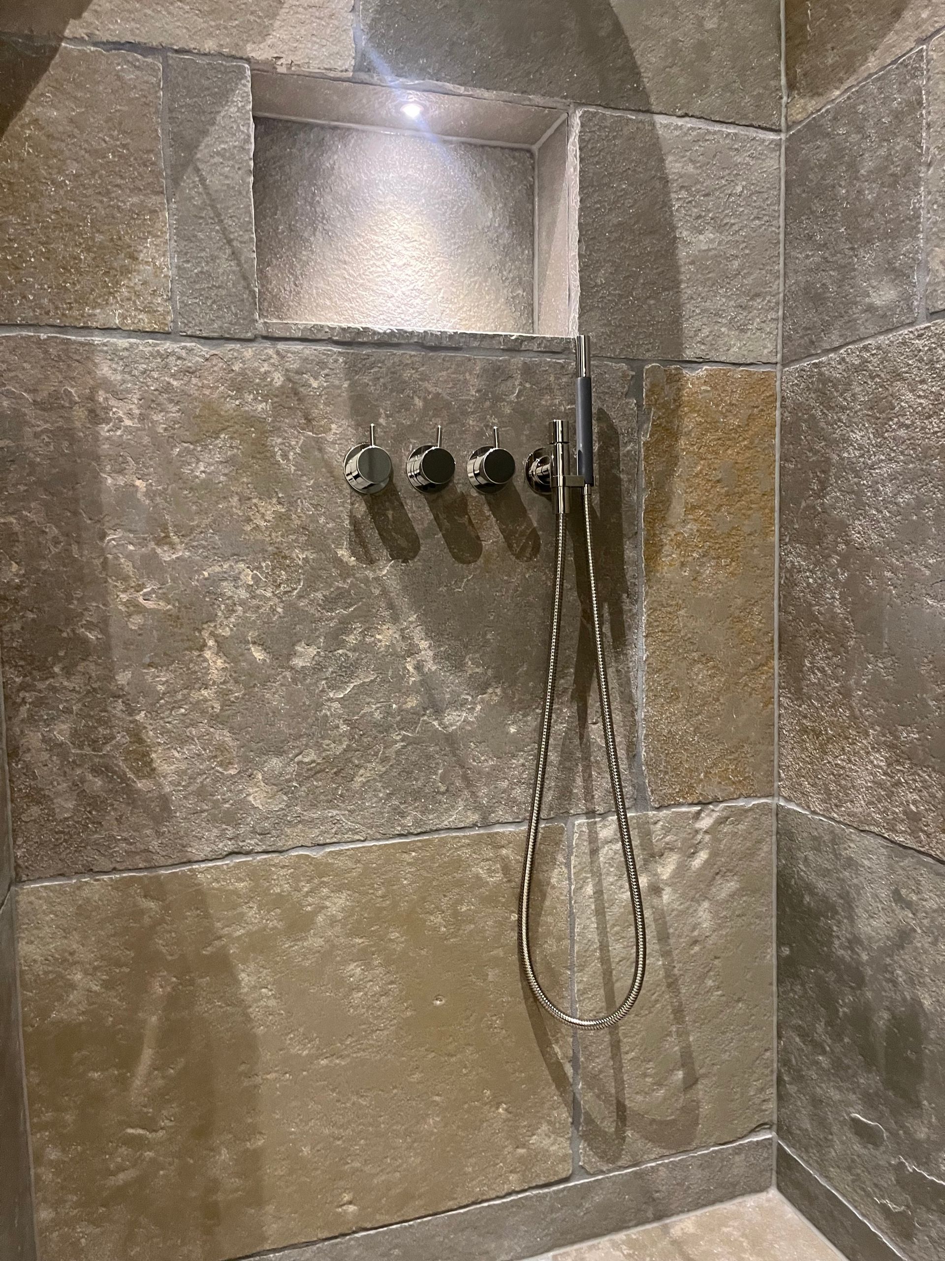 Dusche mit grauen Steinfliesen, Nische, drei Knöpfen und Handbrause.