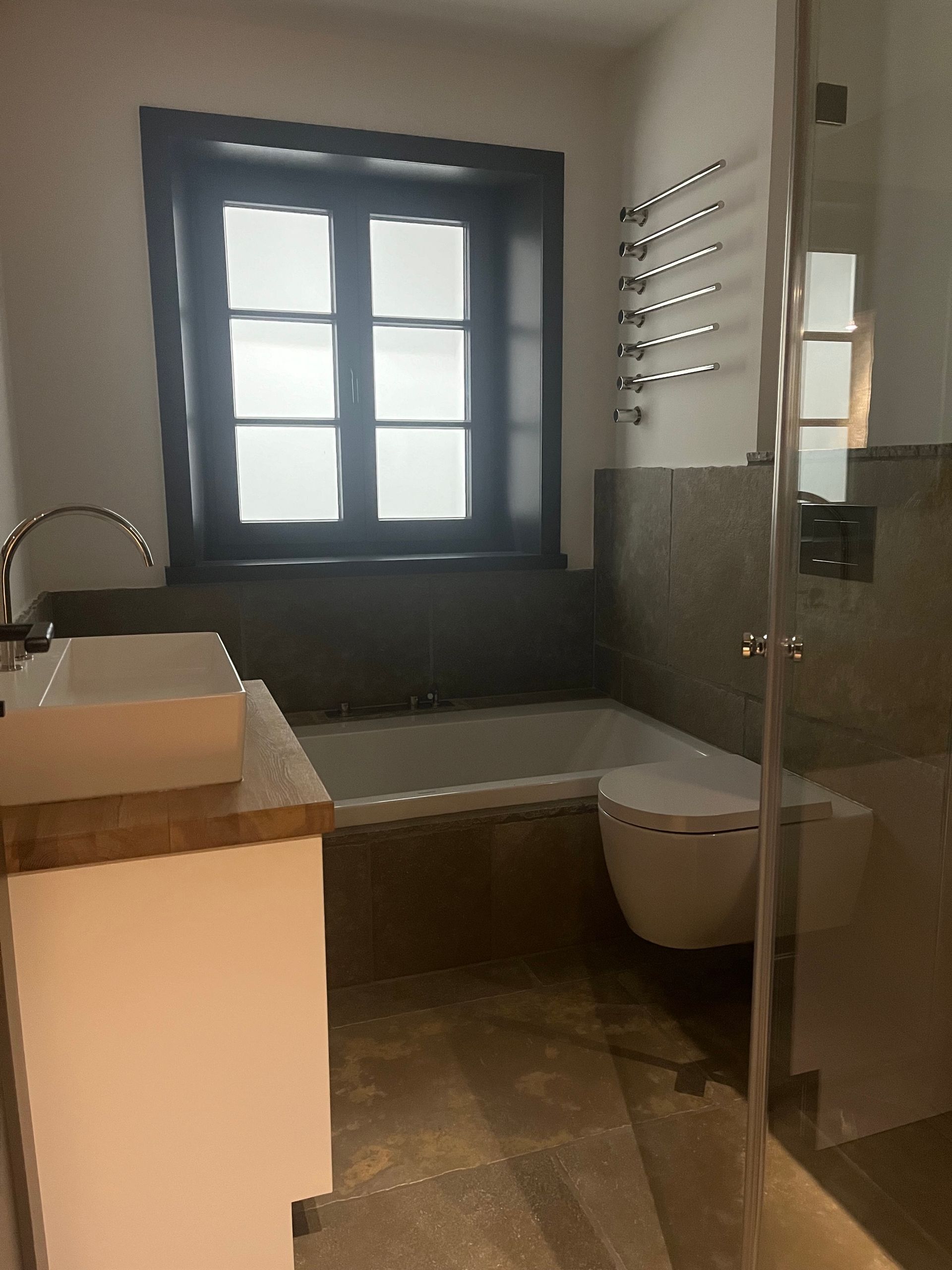 Badezimmer mit Badewanne, Toilette, Waschbecken und Dusche. Dunkler Fensterrahmen, graue Steinwände und ein weißer Waschtisch.