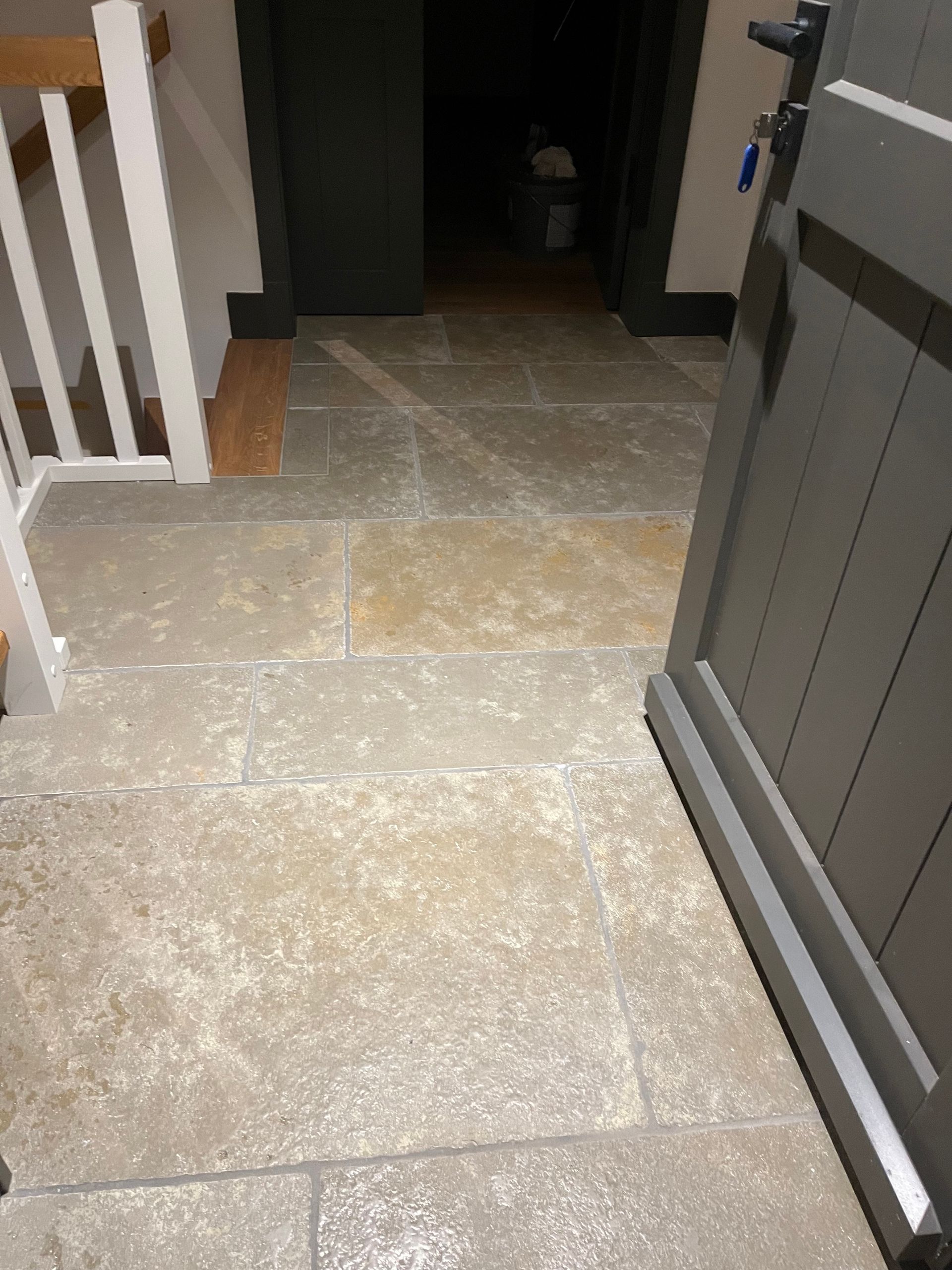 Eingangsbereich mit Steinboden, grauer Tür, Treppe mit weißem Geländer und dunklem Türrahmen.