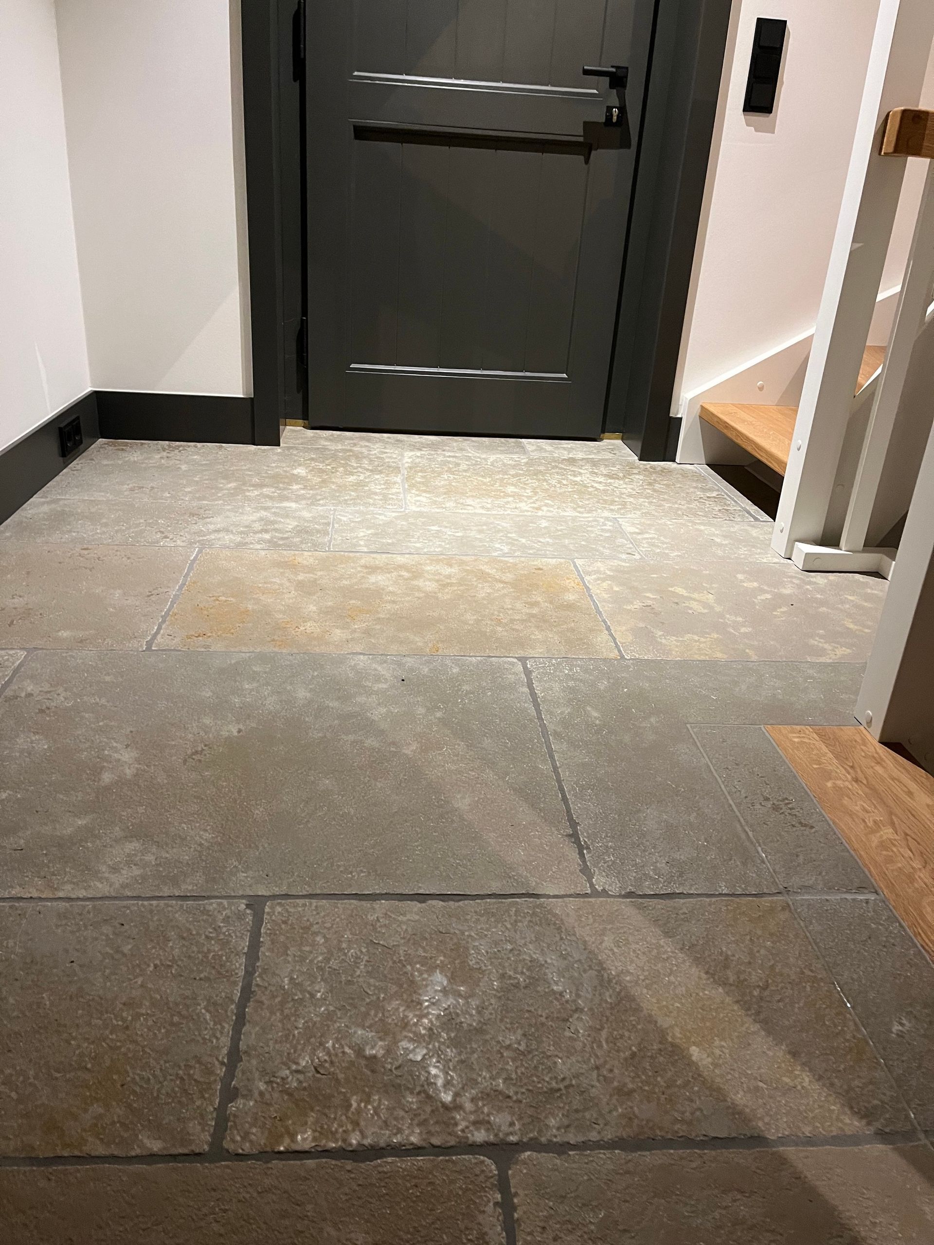 Steinboden in einem Flur mit einer dunkelgrauen Tür und einer Treppe auf der rechten Seite.