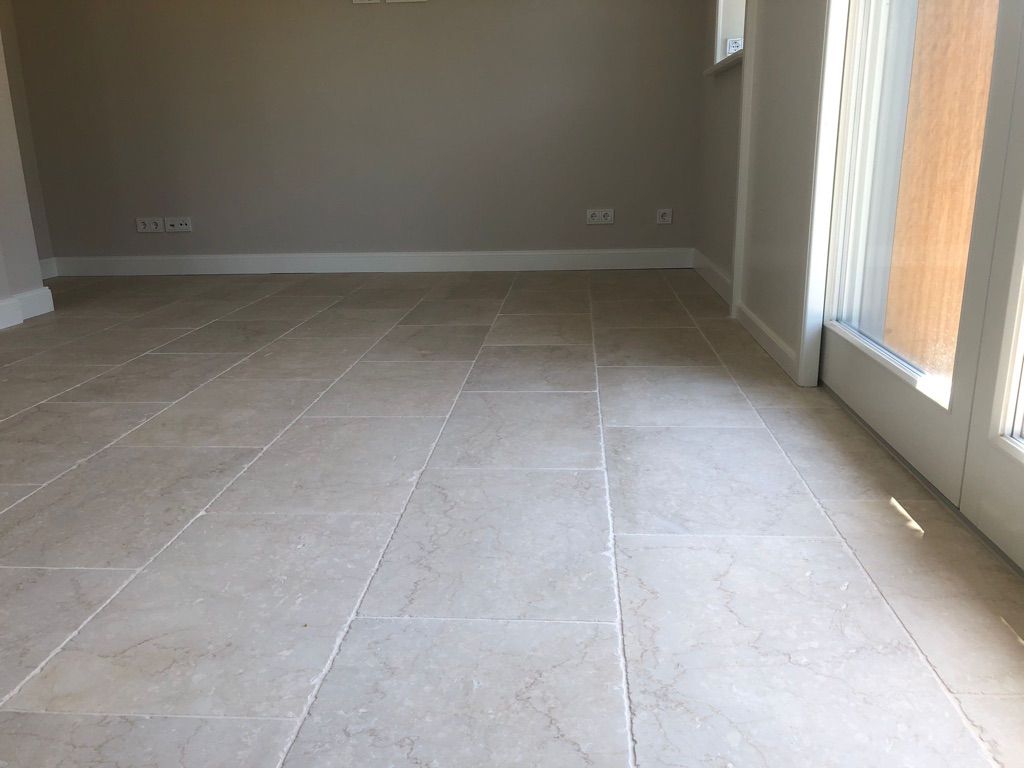 Beiger Fliesenboden in einem Raum mit hellgrauen Wänden und weißen Zierleisten, neben einer Glastür mit weißem Rahmen.