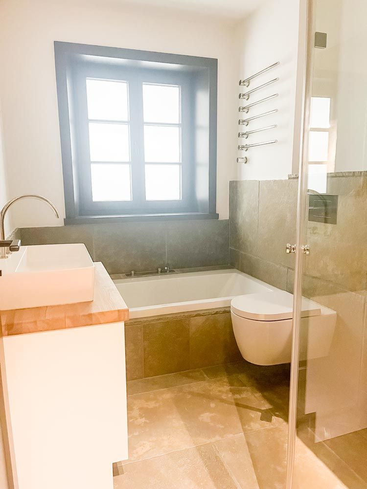 Badezimmer mit Badewanne, Toilette und Waschbecken in der Nähe eines Fensters. Farbschema in Grau und Weiß.