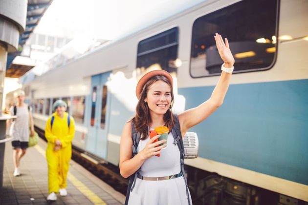 Une femme fait signe de la main sur le quai d'une gare, à côté d'un train bleu et blanc, en tenant une boisson.