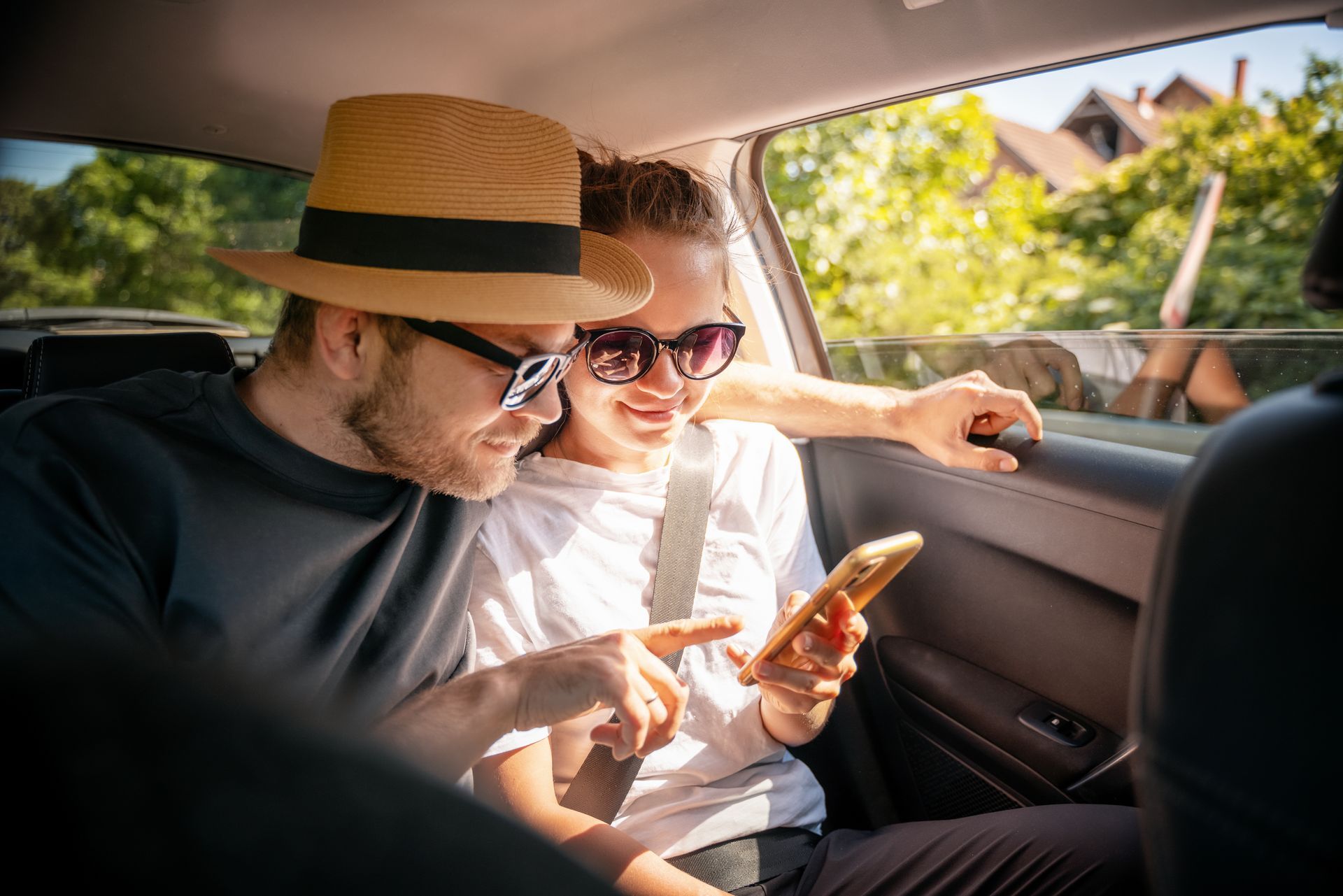 Deux personnes dans une voiture regardent un smartphone, avec le soleil et la verdure à l'extérieur.