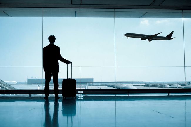 Silhouette d'un voyageur avec sa valise dans un terminal d'aéroport, regardant un avion décoller à travers de grandes fenêtres.