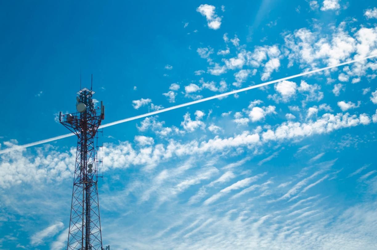 Torre de telefonía celular contra un cielo azul brillante con nubes tenues y una delgada estela blanca.