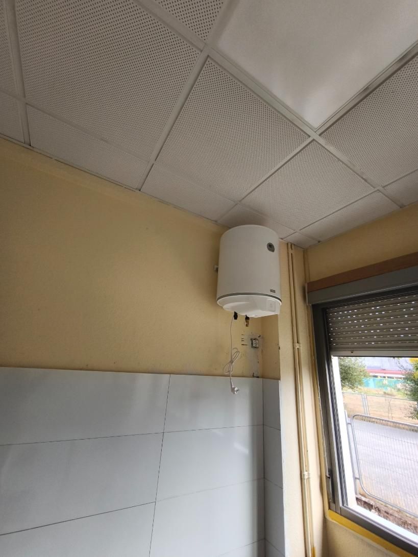 Pequeño rincón de baño alicatado con calentador de agua, paredes beige y una ventana junto a una zona de ducha blanca.