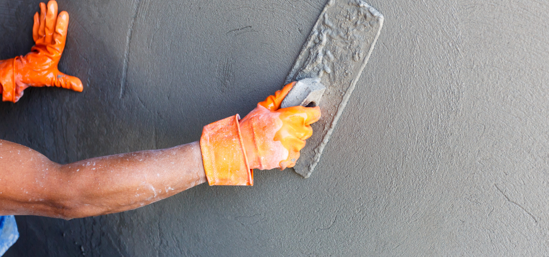 Personne utilisant une truelle pour lisser le béton humide sur un mur, portant des gants orange.