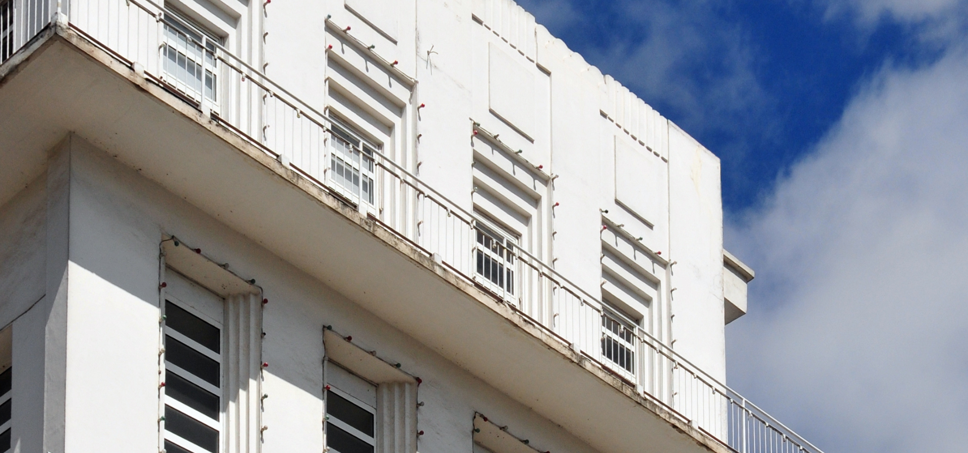 Bâtiment blanc avec balcons et volets horizontaux contre un ciel bleu avec des nuages.