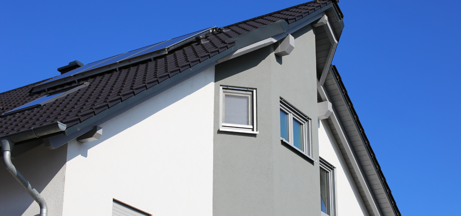 Une maison moderne avec un toit sombre et des murs gris et blancs contre un ciel bleu clair.