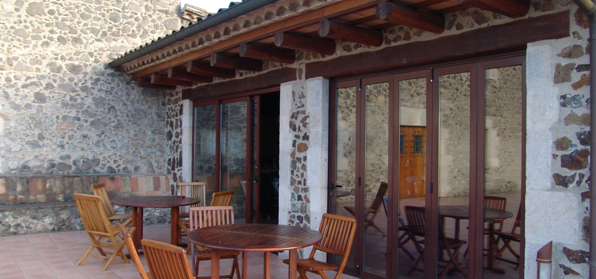 Terrasse extérieure avec tables et chaises en bois, murs en pierre et portes pliantes en verre.