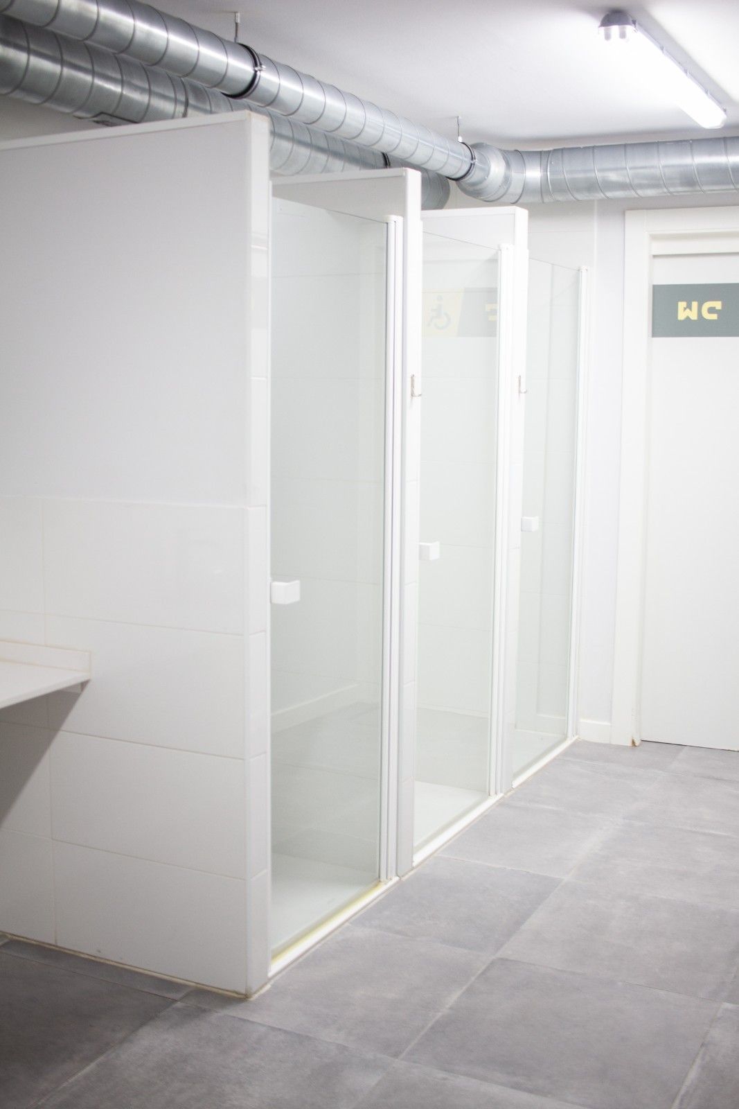Cabinas de baños públicos con mamparas blancas y suelo de baldosas grises bajo conductos de ventilación expuestos.