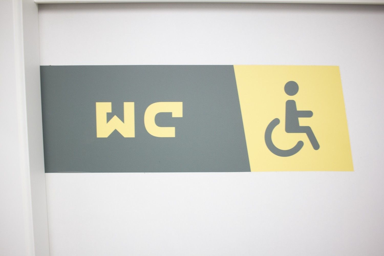 Letrero de baño WC con símbolo de accesibilidad para sillas de ruedas sobre un fondo gris y amarillo.