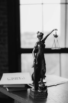 Estatua de la Dama de la Justicia sosteniendo una balanza y una espada sobre un escritorio con libros, frente a una ventana.