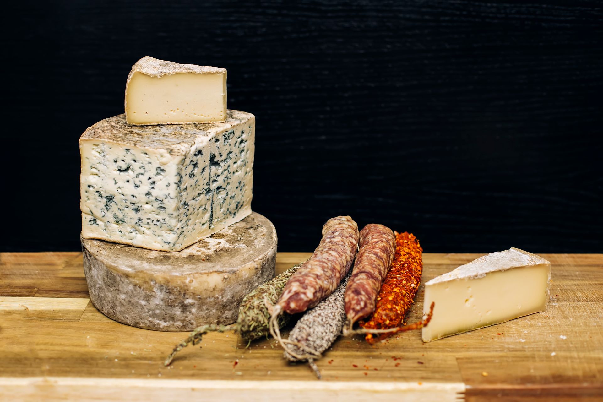 Quesos apilados en una tabla de madera con salchichas sobre un fondo negro.