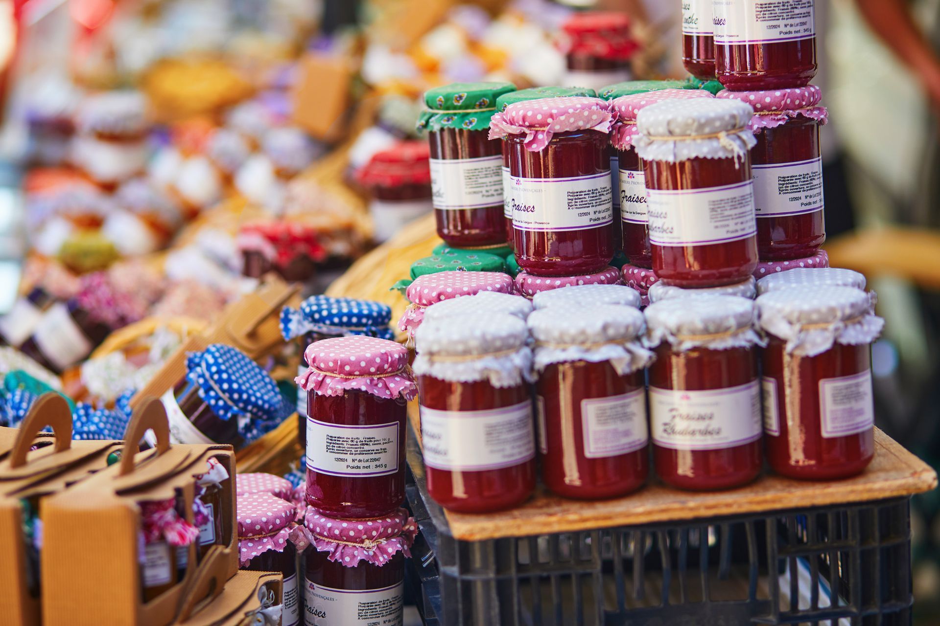 Tarros de mermelada roja con tapas de tela, apilados en una caja de madera en un puesto de mercado.