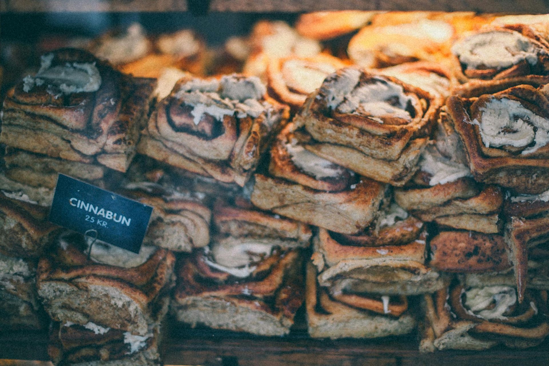 Bollos de canela apilados en la vitrina de una panadería, con un cartel que dice "bollo de canela".