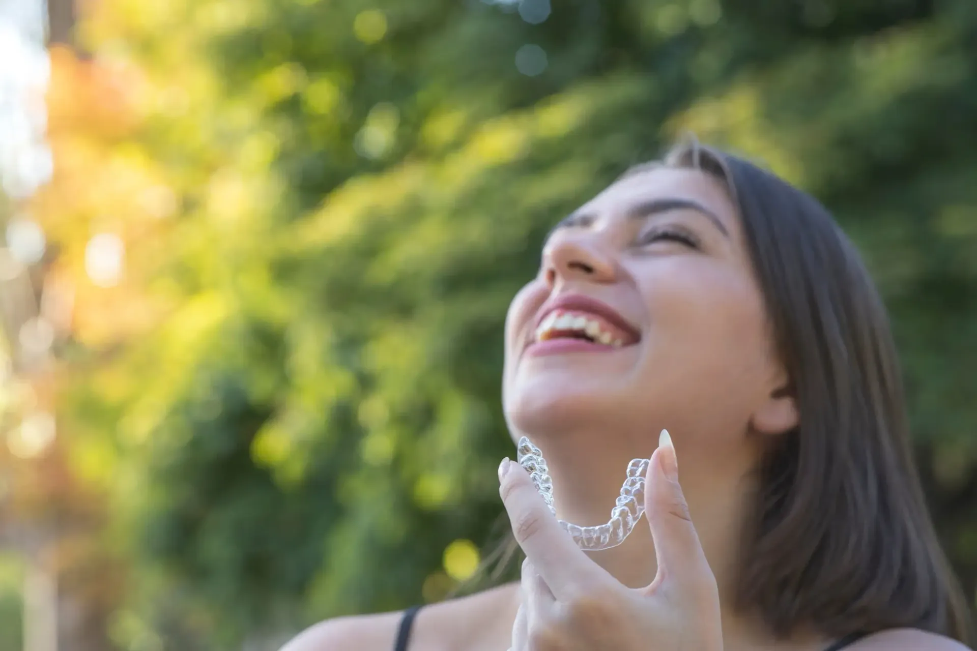 Mujer sonriendo y mirando hacia arriba al aire libre, con la luz del sol y árboles verdes al fondo.