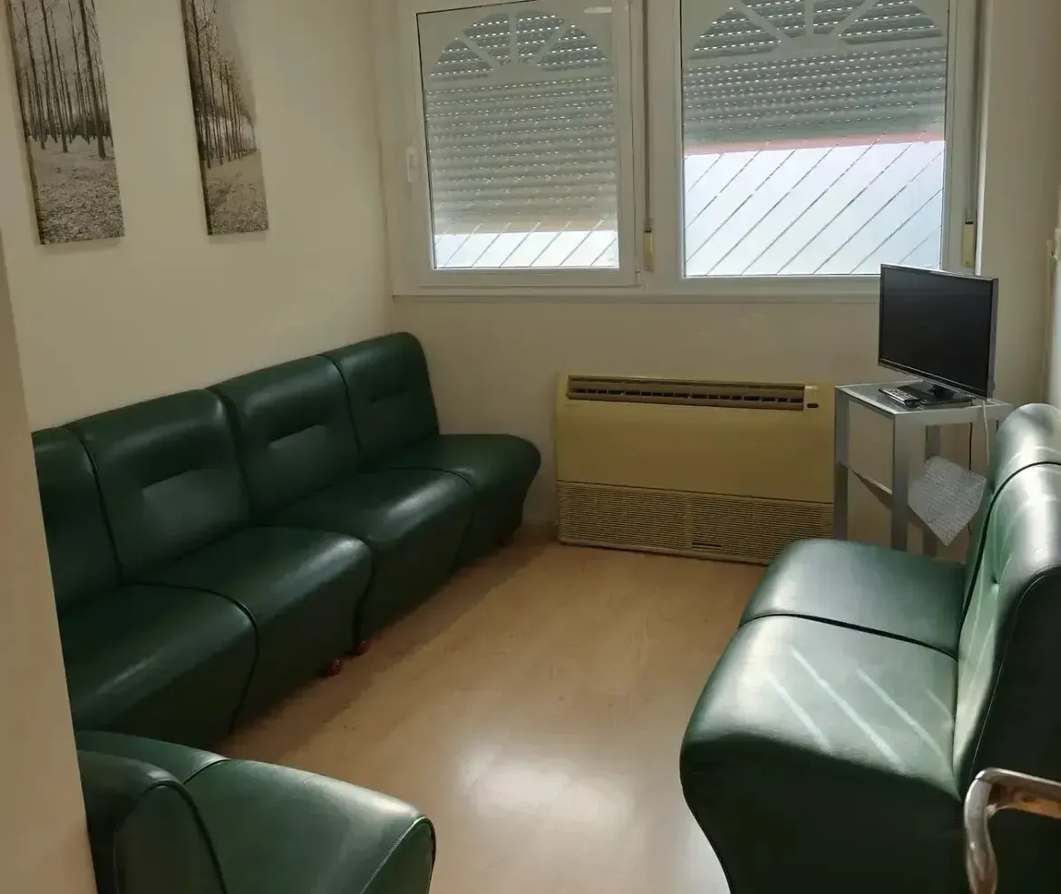 Pequeña sala de estar con sofás de cuero verde, un televisor y ventanas con persianas.