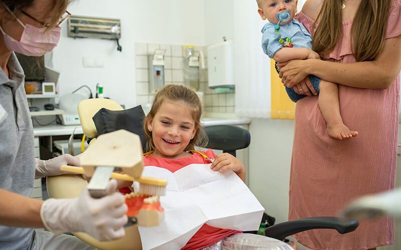 Ein kleines Mädchen sitzt auf einem Zahnarztstuhl, während eine Frau ein Baby hält.