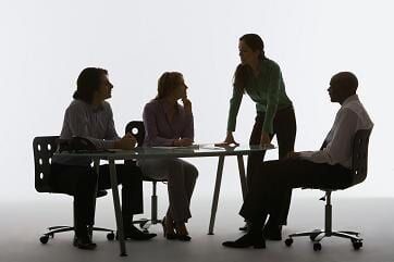Quatro pessoas em silhueta ao redor de uma mesa, uma em pé e as outras sentadas, reunidas em um ambiente interno.