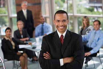 Homem sorridente de terno, com os braços cruzados, em frente a uma reunião de negócios.