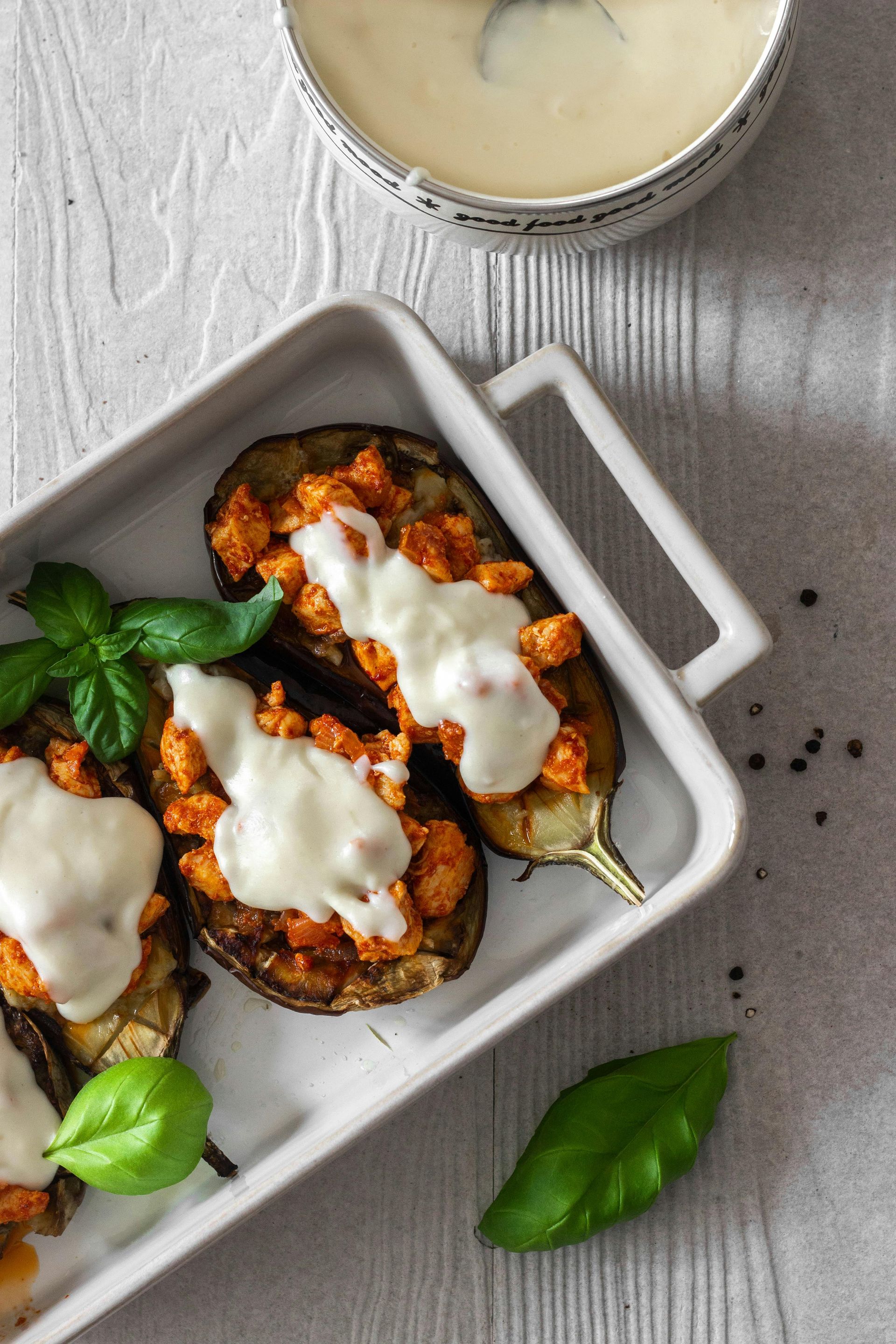 Mitades de berenjena rellenas en un plato blanco, cubiertas con queso, al lado de un bol de salsa y hojas de albahaca.
