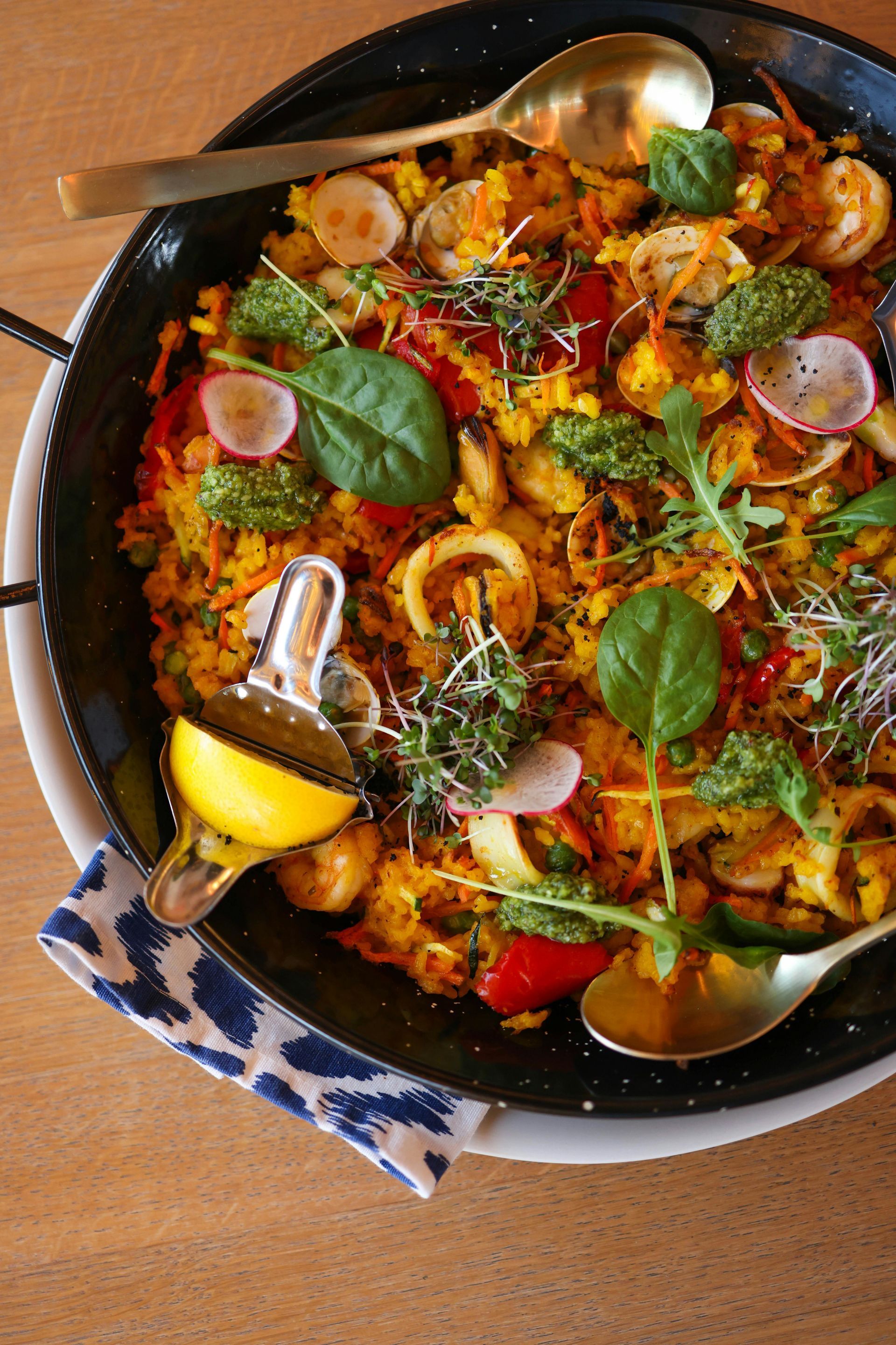 Paella en sartén negra, cubierta con verduras, hierbas y limón.